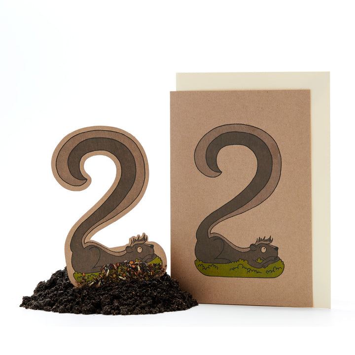 Deux sculptures en papier brun ressemblant au chiffre 2, une sur papier cartonné et une sur terre, avec des détails de dessin ressemblant à des dragons endormis.