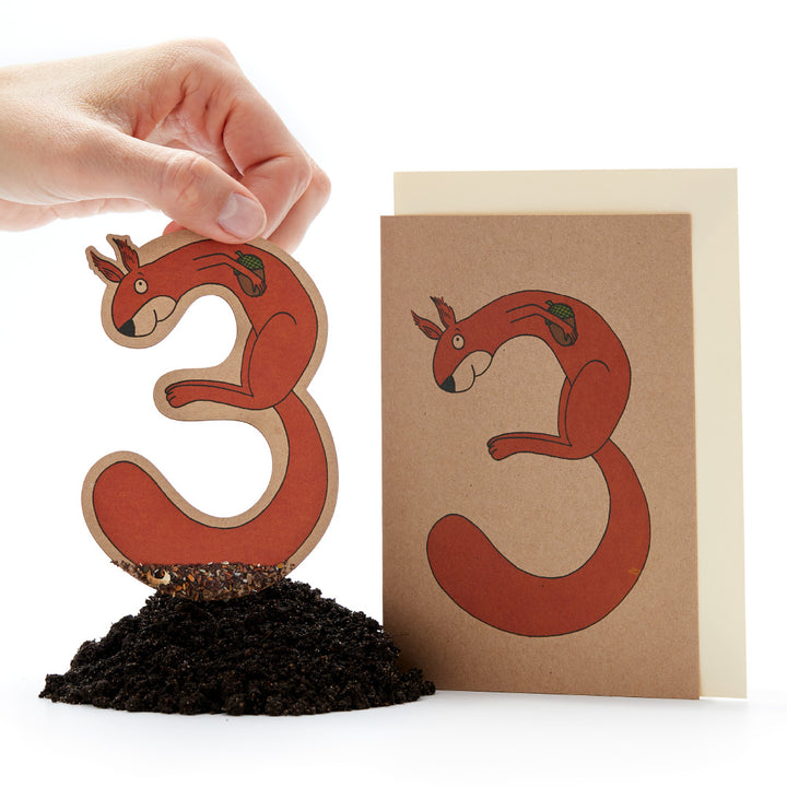 Main tenant une figurine de plante d'écureuil roux à côté d'une carte de vœux avec le même motif sur fond blanc.
