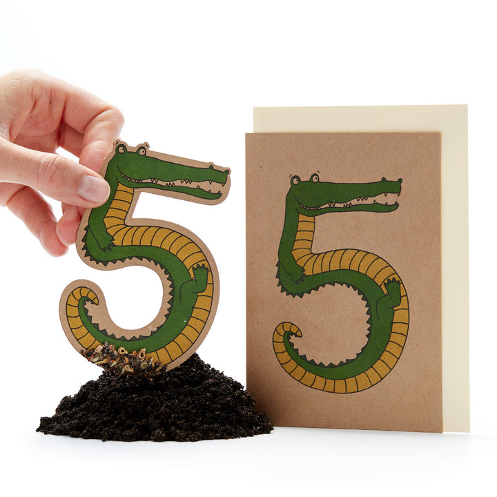 Main tenant une figurine en papier découpée en forme de « 5 » qui ressemble à un crocodile, à côté d'une carte avec le même motif sur terre.