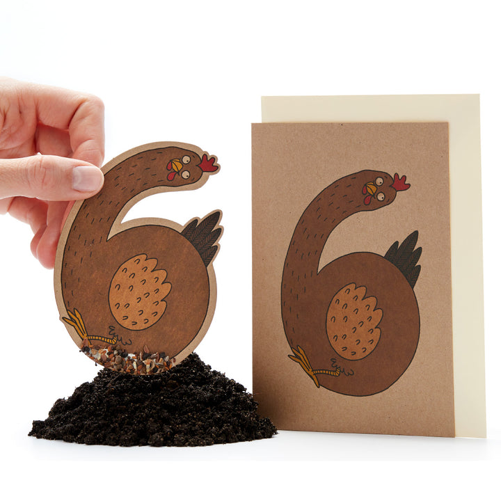 Une main tient une découpe en papier représentant un poulet en forme de 6, à côté d'une carte avec la même image et un tas de terre au fond.
