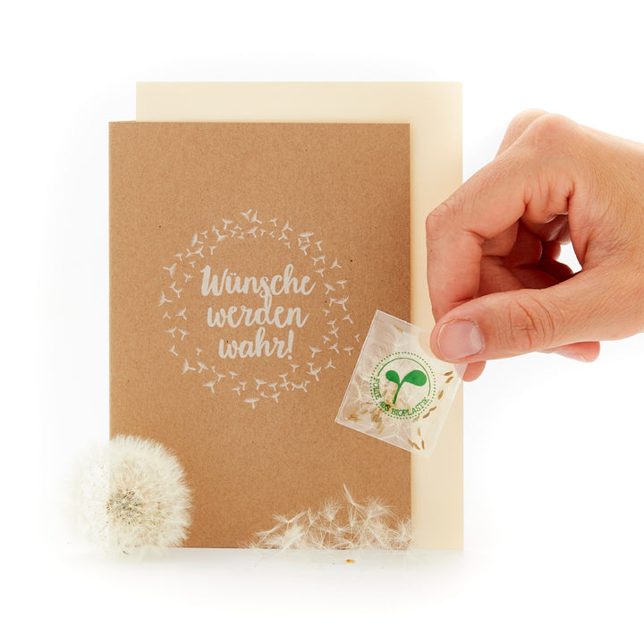 Une main tient une carte biodégradable avec l'inscription « Les vœux se réalisent ! », entourée de pissenlits.