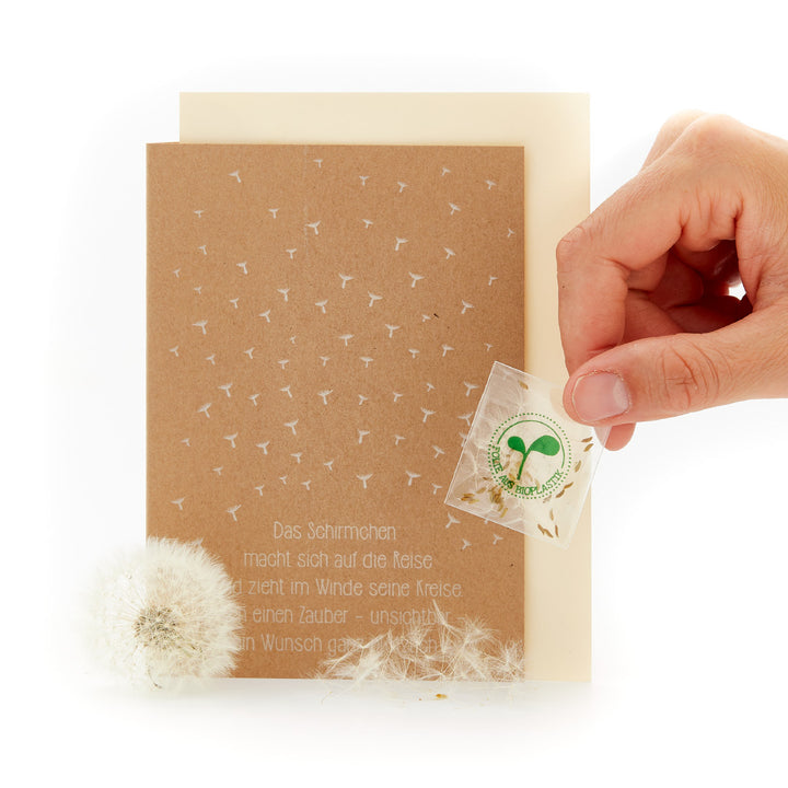 Main tenant une carte de vœux avec des graines de pissenlit et un poème à côté d'un pissenlit.