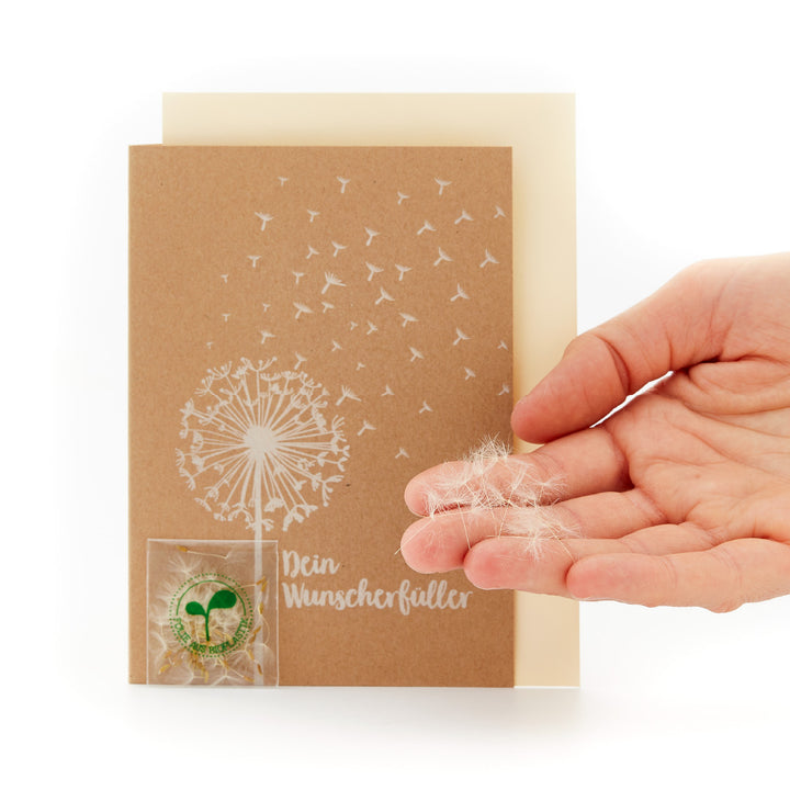 Main tenant une carte avec un motif de pissenlit et l'inscription « Votre exauceur de souhaits » et un petit sac transparent contenant des graines de pissenlit.
