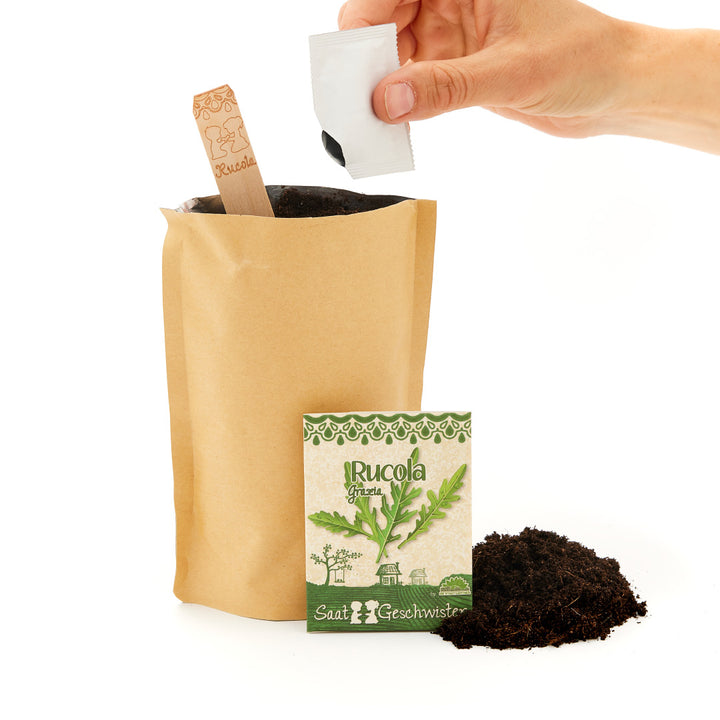 Une main ouvre un paquet de graines de roquette à côté d'un sac en papier contenant de la terre et d'un panneau en bois sur lequel est écrit « Roquette ».