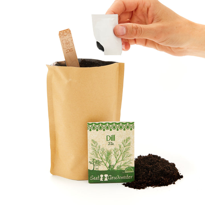 Main versant des graines dans un sac en papier, à côté d'un paquet de graines d'aneth et d'une pelle de terre.