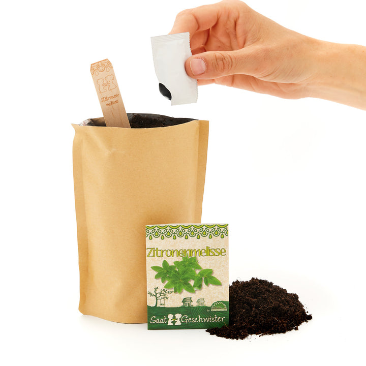 Une main ouvre un sachet de graines sur un sac de plantation marron contenant de la terre et une pancarte indiquant « Mélisse »