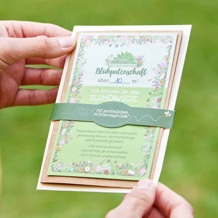 Deux mains tiennent un certificat de parrainage de fleurs sur fond vert, avec un cadre floral et un texte pour le cadeau d'une prairie fleurie.