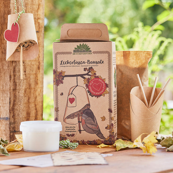 Emballage d'un kit de friandises pour oiseaux avec cloche d'alimentation et décoration naturelle en arrière-plan.