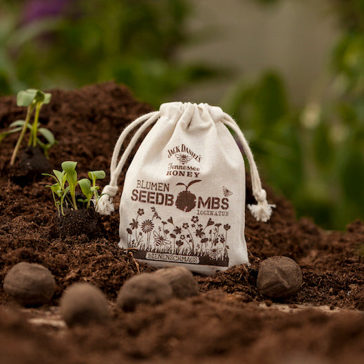 Un petit sac en tissu étiqueté « Bombes de graines de fleurs » est posé sur un sol fertile à côté des semis et des bombes de graines.