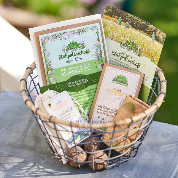 Un panier en fil de fer contenant divers produits et dépliants sur le parrainage de fleurs et des graines de fleurs sauvages de « Die Stadtgärtner » (Les jardiniers de la ville) est posé sur une table à l'extérieur, à la campagne.