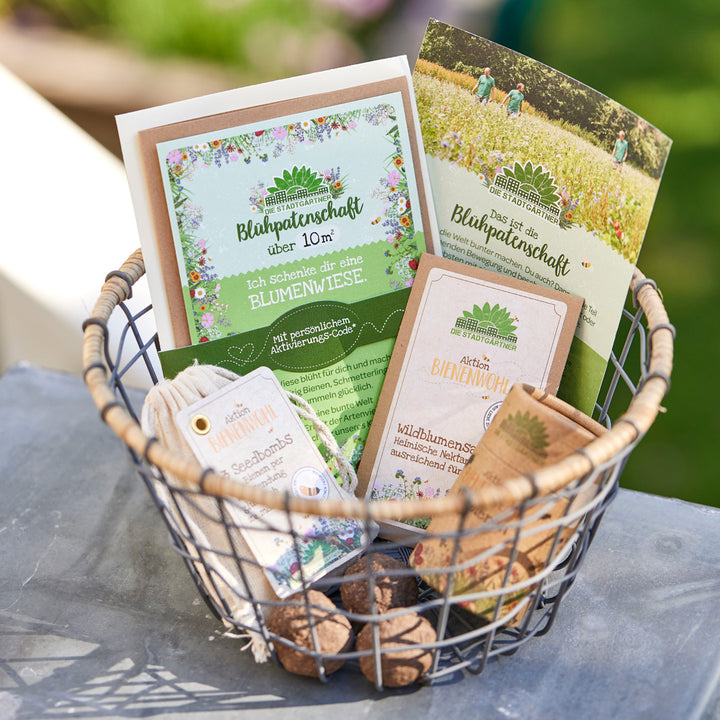 Un panier en fil de fer contenant divers produits et dépliants sur le parrainage de fleurs et des graines de fleurs sauvages de « Die Stadtgärtner » (Les jardiniers de la ville) est posé sur une table à l'extérieur, à la campagne.